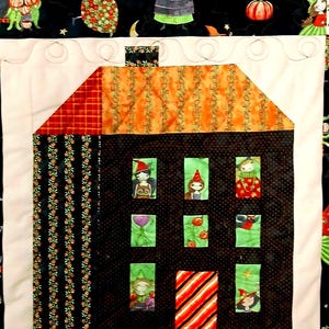 May include: A quilted wall hanging featuring a central house block with a black polka-dot facade, orange roof, and striped door. The house has six green windows containing small figures. The border is black fabric with a colorful Halloween print.