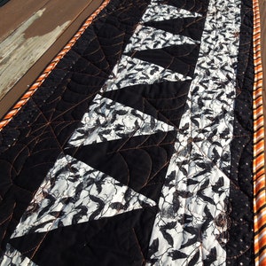 May include: Black and white table runner with a bat pattern and orange trim. The runner is quilted with a spiderweb design.