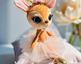 Op de afbeelding: Een pluche hertenknuffel met grote donkere ogen, gekleed in een lichtroze tule tutu en gouden balletschoentjes. Het hert heeft een beige vacht met witte stippen, roze gevoerde oren en een klein roze bloemetje tussen twee gouden geweien.