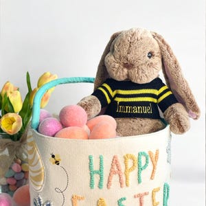 May include: A white canvas Easter basket with a blue handle, filled with pink and orange Easter eggs. A brown bunny wearing a black and yellow striped sweater with the name "Immanuel" embroidered on it is sitting in the basket. The basket is decorated with embroidered flowers and the words "Happy Easter".