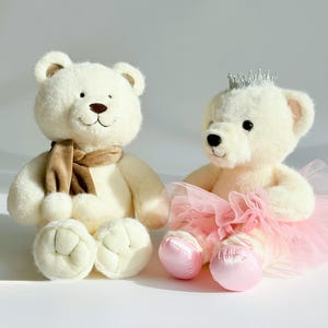 May include: Two plush teddy bears. One bear wears a brown scarf, the other a silver crown and pink tutu. Both are cream-colored with brown eyes and noses. The bears have stitched smiles and rounded paws, perfect for a child's toy or nursery decor.