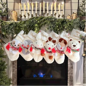 Op de afbeelding: Een rij van zes cr&egrave;mekleurige pluche kerstsokken die op een rij hangen. Elke sok is voorzien van een ander knuffeldier met een rood, groen en wit patroon sjaaltje. De namen Max, Taylor, Sophie, Dad, Mom en Baby 1st Christmas zijn in gouddraad op de boorden geborduurd.