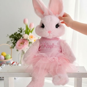 May include: A plush pink bunny wearing a pink tutu and a top with the name "Kayla" embroidered on it. The bunny has long ears, black eyelashes, and is sitting on a white bench. A hand is gently touching the bunny's ear. Easter themed.