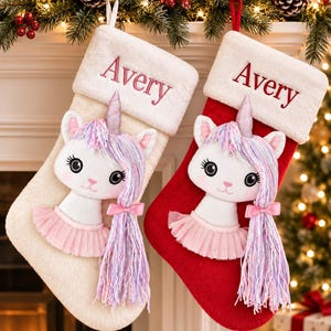 Op de afbeelding: Twee pluche kerstsokken, &eacute;&eacute;n cr&egrave;me en &eacute;&eacute;n rood, elk met een witte rand van imitatiebont geborduurd met de naam Avery in het rood. Een witte eenhoornapplicatie met een roze tutu, een zilveren hoorn en lang haar van pastelkleurig garen.