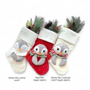Op de afbeelding: Drie kerstsokken met pluche pingu&iuml;napplicaties. E&eacute;n is wit met een witte sjaal, &eacute;&eacute;n is rood met een sjaal met patroon, en &eacute;&eacute;n is wit met een sjaal met patroon. Elke sok heeft een witte rand van imitatiebont en dennentakken.