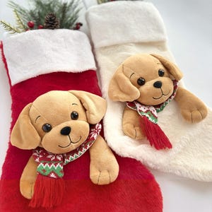 Op de afbeelding: Twee pluche kerstsokken, &eacute;&eacute;n rood en &eacute;&eacute;n cr&egrave;me, elk met een zachte, beige puppy aan de voorzijde bevestigd. Elke puppy draagt een kleine, gebreide sjaal met een rood, groen en wit geometrisch patroon en heeft donkere ogen.
