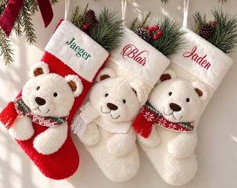 Op de afbeelding: Drie kerstsokken met pluche witte berengezichten en sjaals. De linker sok is rood met de naam Jaeger, de middelste is wit met Blair en de rechter is wit met Jaden. Elke sok heeft een witte boord en is gevuld met dennentakken.