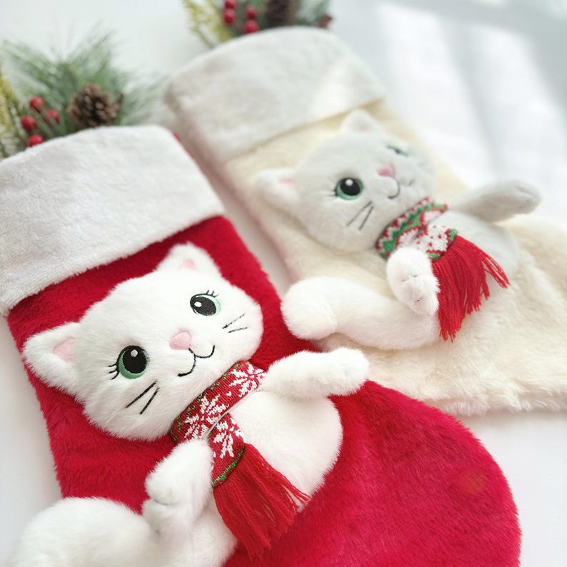 Op de afbeelding: Twee pluche kerstsokken, &eacute;&eacute;n rood en &eacute;&eacute;n cr&egrave;me, elk voorzien van een zachte witte kat met groene ogen en een rood-witte sjaal met patroon. Beide sokken hebben een witte rand van imitatiebont aan de bovenkant en een textuurrijke, pluizige stof.