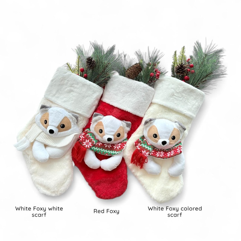 Op de afbeelding: Drie pluche kerstsokken met vossengezichten en sjaals. De linker is wit met een witte sjaal, de middelste is rood met een sjaal met patroon, en de rechter is wit met een sjaal met patroon. Elke sok bevat dennentakken en dennenappels.