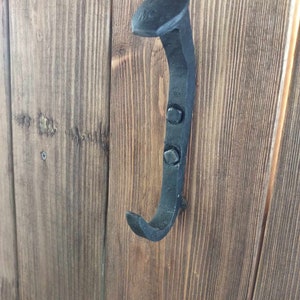 May include: A black metal hook made from a repurposed nail. The hook is mounted on a wooden surface.