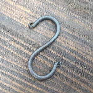 Könnte beinhalten: Ein einzelner, handgeschmiedeter S-förmiger Haken aus Metall. Der Haken ist dunkelgrau und hat eine glatte, leicht gekrümmte Oberfläche.