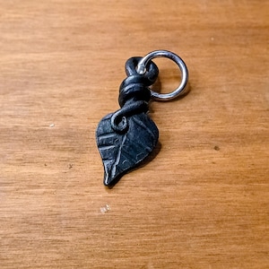 May include: A dark gray, hand-forged metal pendant shaped like a leaf with a textured surface and a coiled stem. A polished silver-colored metal jump ring is attached to the top of the coiled stem, allowing the piece to be suspended as a charm.