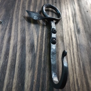 May include: A black metal hook with a leaf design. The hook is attached to a wooden surface.
