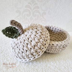 May include: A beige crochet apple-shaped container with a removable lid. The apple features a textured stitch pattern, a light brown stem, and a dark green leaf attached to the top. The text Mrs. KaWi Handmade is visible in the bottom left corner.