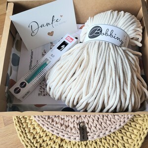 May include: A cardboard box containing a large skein of off-white Bobbiny cotton cord, a Prym Ergonomics 8.0 mm crochet hook in its packaging, a white card with the text Danke Sch&ouml;n, and a printed instruction sheet for a coaster project.