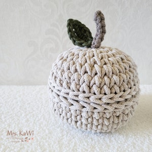 May include: A round, beige crochet container shaped like an apple with a textured, braided stitch pattern. It features a dark green crocheted leaf and a light brown crocheted stem on top. The text Mrs. KaWi Handmade is visible in the bottom left corner.