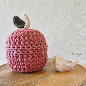 May include: A round, dusty rose crochet container with a lid, featuring a dark green leaf and a beige stem on top. Beside it sits a small, smooth, light pink ceramic bird figurine. The text Mrs. KaWi is visible in the lower right corner.
