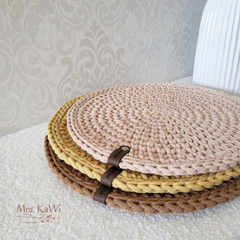 May include: Three round, crocheted mats stacked vertically in shades of light pink, mustard yellow, and brown. Each mat features a thick, textured cord construction and a small, dark brown faux leather tag labeled Mrs. KaWi attached to the edge.