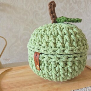 May include: A light green crocheted apple-shaped container with a brown stem and a green leaf. The container features a brown leather-like tab with the text Mrs. Kawi embossed on it, secured by a small gold-colored metal rivet on the side.