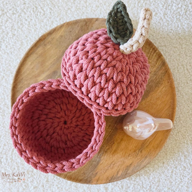 May include: A round, crocheted container shaped like an apple in a dusty rose color. The lid features a dark green crocheted leaf and a light beige stem. The interior of the container is hollow and shows the same dusty rose yarn texture.