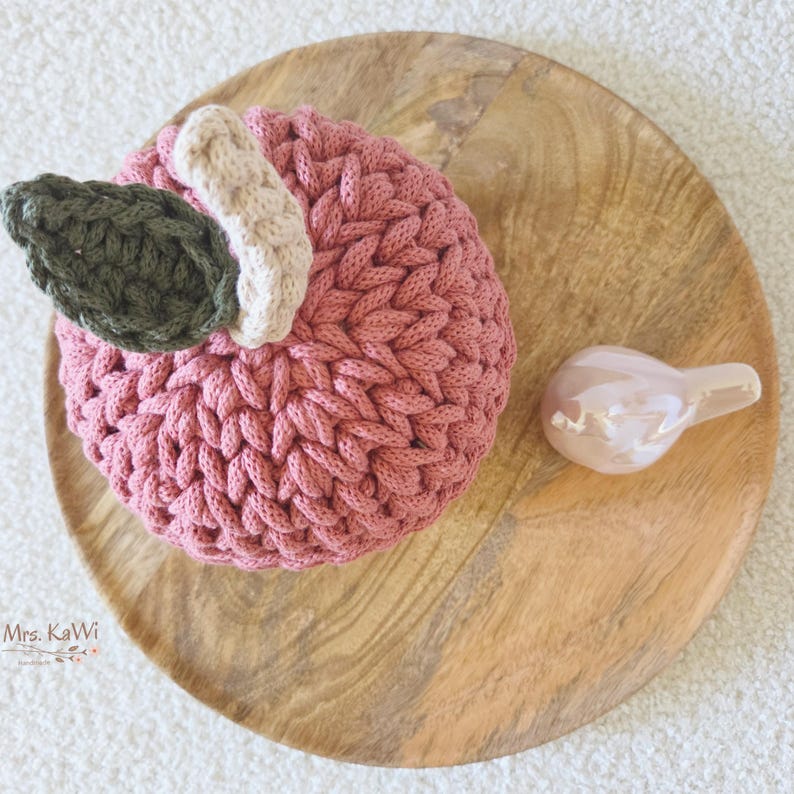 May include: A round, crocheted object in a dusty rose color with a thick, textured yarn construction. It features a small, cream-colored stem and a single, dark olive green crocheted leaf attached to the top. The text Mrs. KaWi is visible.