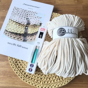 May include: A craft kit featuring a skein of off-white Bobbiny cord, a Prym 8.0 mm crochet hook in its packaging, and a printed instruction sheet titled "Platzset/Untersetzer" by Mrs. KaWi handmade, resting on a round mustard yellow crocheted mat.