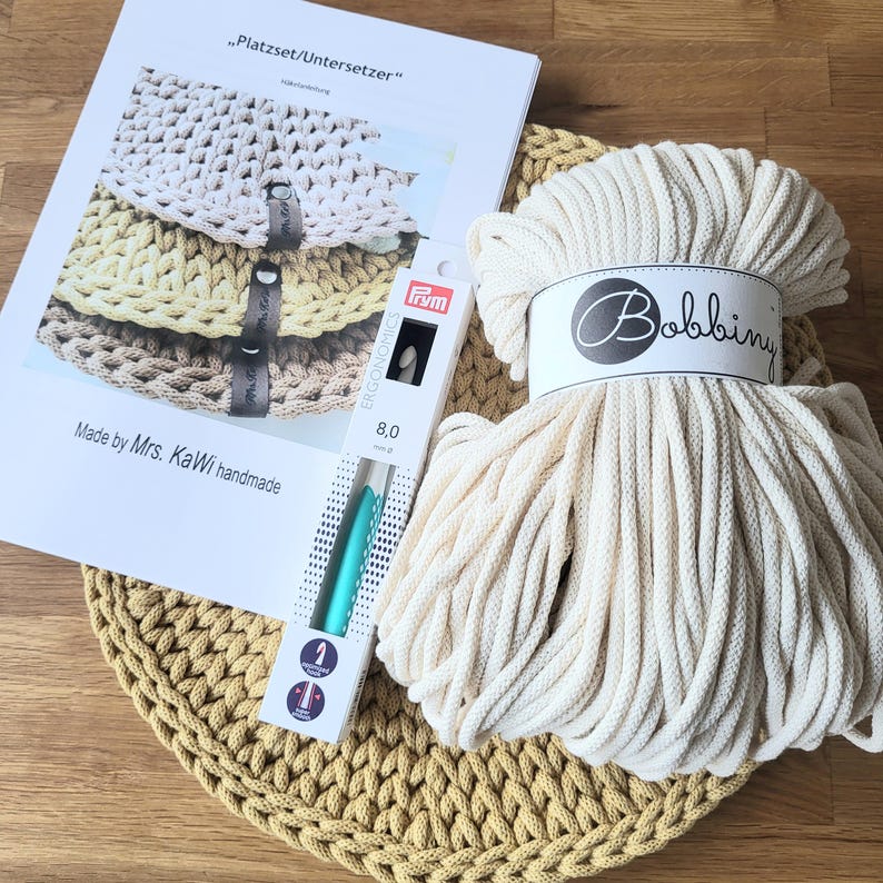 May include: A craft kit featuring a skein of off-white Bobbiny cord, a Prym 8.0 mm crochet hook in its packaging, and a printed instruction sheet titled "Platzset/Untersetzer" by Mrs. KaWi handmade, resting on a round mustard yellow crocheted mat.