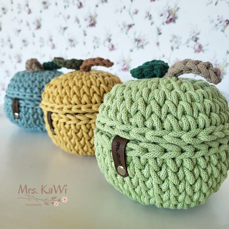 May include: Three round, crocheted storage containers shaped like apples in light green, mustard yellow, and dusty blue. Each container features a dark green crocheted leaf, a braided brown stem, and a brown leather tab with the text Mrs. KaWi.