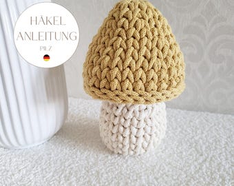 May include: A crochet mushroom figure with a mustard yellow cap and a cream-colored stem. A circular white label is visible with the text HÄKEL ANLEITUNG PILZ and a small German flag icon. The mushroom has a thick, textured yarn construction.