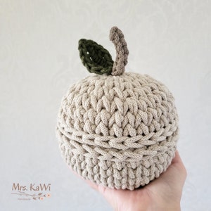 May include: A round, beige crocheted apple-shaped container with a lid. The top features a dark green crocheted leaf and a light brown crocheted stem. The item is constructed from thick, textured yarn with visible stitches throughout the surface.