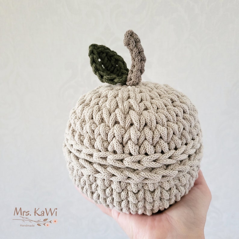 May include: A round, beige crocheted apple-shaped container with a lid. The top features a dark green crocheted leaf and a light brown crocheted stem. The item is constructed from thick, textured yarn with visible stitches throughout the surface.