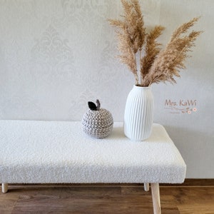 May include: A beige crocheted apple-shaped container with a dark brown leaf and stem, placed next to a white ribbed ceramic vase filled with dried pampas grass. The text Mrs. KaWi is visible on the right side of the image near the vase.