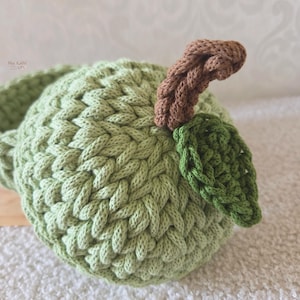 May include: A light green crocheted apple with a brown stem and a dark green leaf. The apple features a textured, braided stitch pattern throughout its round body. The small text "Mes KaWi" is visible in the upper left corner of the image.