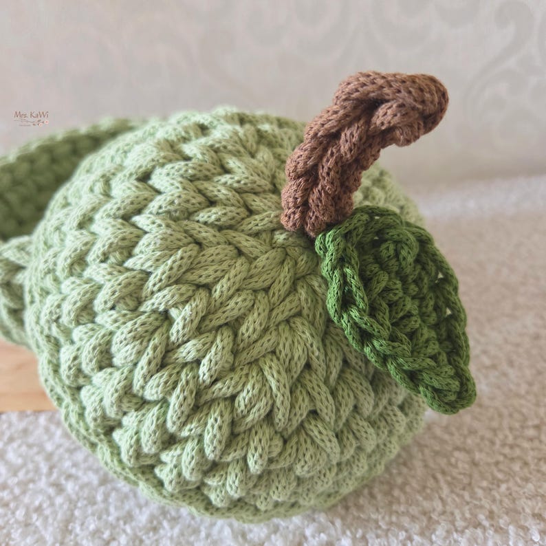 May include: A light green crocheted apple with a brown stem and a dark green leaf. The apple features a textured, braided stitch pattern throughout its round body. The small text "Mes KaWi" is visible in the upper left corner of the image.