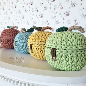 Pode incluir: Quatro cestos de armazenamento redondos de crochê em forma de maçã nas cores ferrugem, azul claro, amarelo e verde claro. Cada cesto apresenta um pequeno caule e folha de crochê no topo, com uma etiqueta de couro marrom Mrs. KaWi na lateral.