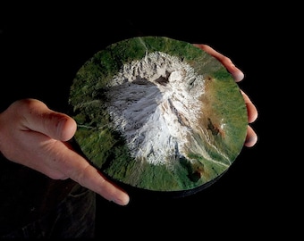 May include: A 3D model of a mountain with a snow-capped peak, surrounded by green vegetation. The model is held in a person's hands.