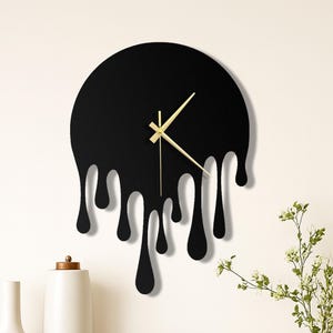 May include: A black wall clock with a unique melting design. The clock face is a black circle with gold-colored hands. The bottom of the clock has a dripping effect. The clock is mounted on a white wall.