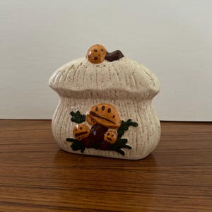 May include: A ceramic mushroom-shaped container with a textured, off-white finish. The container is decorated with brown and orange mushroom designs and green leaves. The lid has a small mushroom on top.