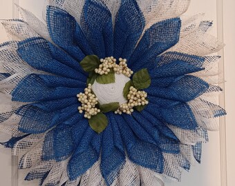 May include: A handcrafted wreath with a flower design. The wreath features layers of blue and white mesh petals, arranged in a circular pattern. The center has a white woven base with small white berries and green leaves. The wreath is designed for door or wall decor.