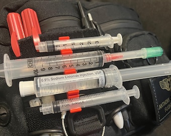 May include: A black medical bag with a variety of medical supplies, including syringes, a saline solution bottle, and a red and white first aid kit. The bag has a patch that says "Anthony Flight Paramedic".
