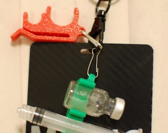 May include: A black badge with the word "MED" in white, featuring a syringe, a vial, and a red plastic accessory. The badge is attached to a black lanyard with a metal clasp and ring.