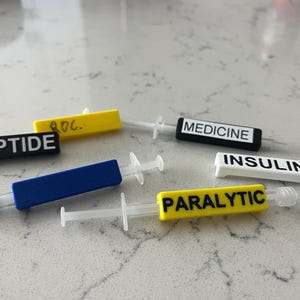 May include: Several syringes with colorful labels. The labels read "PEPTIDE", "MEDICINE", "INSULIN", and "PARALYTIC". The syringes have clear barrels and white plungers. The labels are black, white, yellow, and blue.