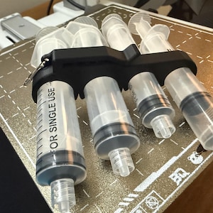 May include: A black plastic holder securing four clear plastic syringes of varying sizes. Each syringe features black measurement markings and the text FOR SINGLE USE printed on the side. The holder is designed to keep the syringes organized.