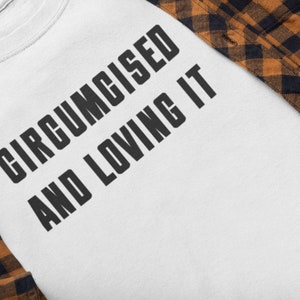 May include: White t-shirt with the text "CIRCUMCISED AND LOVING IT" in bold, black letters. A brown and black plaid flannel shirt partially covers the t-shirt.