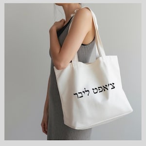 May include: Cream-colored tote bag with black Hebrew text. The bag is carried over the shoulder. The text on the bag reads "צ'אפט ליבר". The bag is made of a soft, durable material.