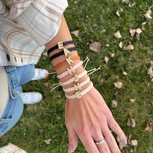 May include: A person's wrist wearing several friendship bracelets with different coloured cords and gold letter charms. The charms spell out the word "EAKHE"