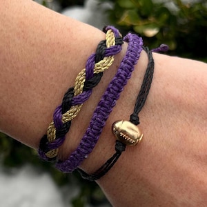 May include: Three friendship bracelets in purple, black, and gold with a gold football charm. The bracelets are stacked on a person's wrist.