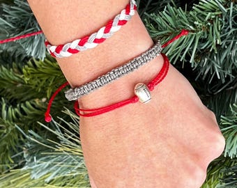 May include: Three bracelets on a wrist. The top bracelet is braided with red, white, and gray string. The middle bracelet is gray and the bottom bracelet is red with a silver football charm.