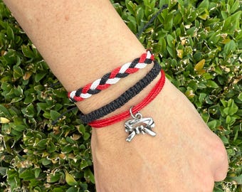 May include: A set of three braided bracelets in red, black, and white. One bracelet features a silver-colored karate belt charm. The bracelets are worn on a wrist against a backdrop of green foliage.