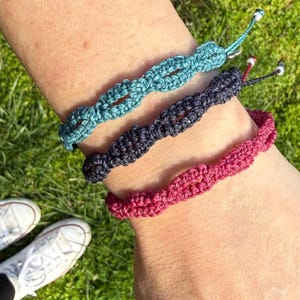 May include: Three handmade macrame bracelets in teal, navy, and magenta. Each bracelet features a woven design and adjustable closure with a small silver bead. The bracelets are displayed on a wrist against a background of green grass.